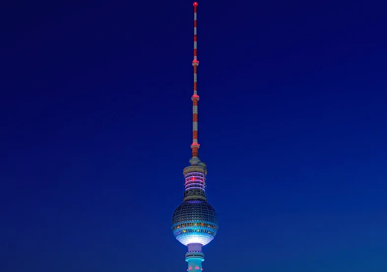 A tall, illuminated television tower with a spherical middle section and a red-and-white striped antenna against a dark blue…