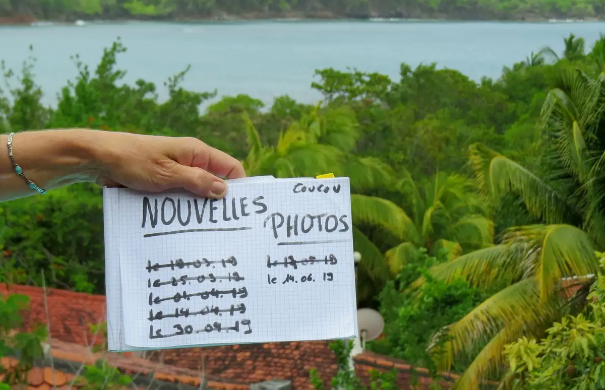 A hand holds a handwritten note on graph paper against a tropical backdrop of palm trees and ocean. The note, written in…