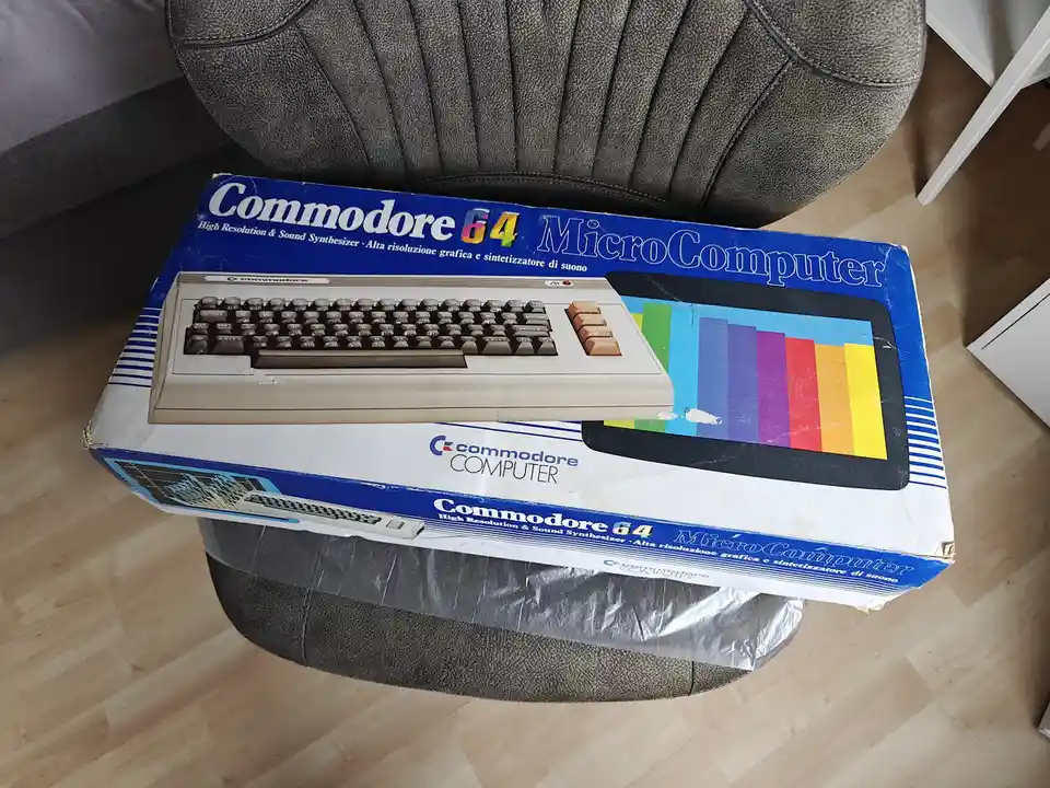 A weathered blue and white box for a Commodore 64 MicroComputer sits on a gray upholstered chair, showing visible wear and…