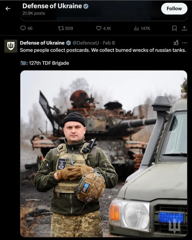 A Ukrainian soldier in full combat gear stands in front of a destroyed, burnt-out Russian tank, holding a helmet. A military…