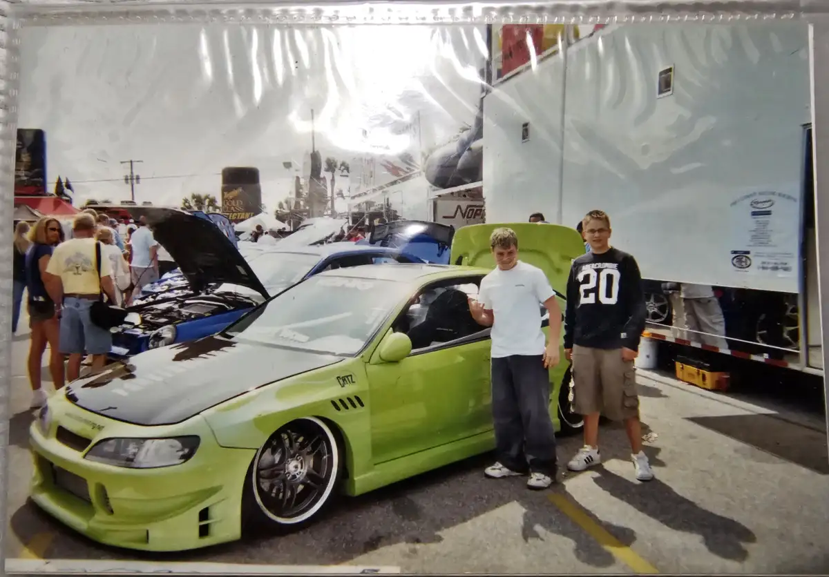 Two young men stand beside a lime-green modified car with its hood open at a car show, surrounded by other vehicles and…