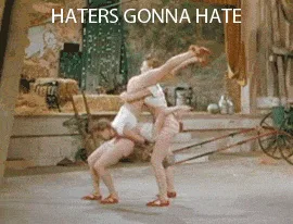 A six-frame GIF shows two women in white outfits performing a synchronized, acrobatic dance move in a rustic barn setting…