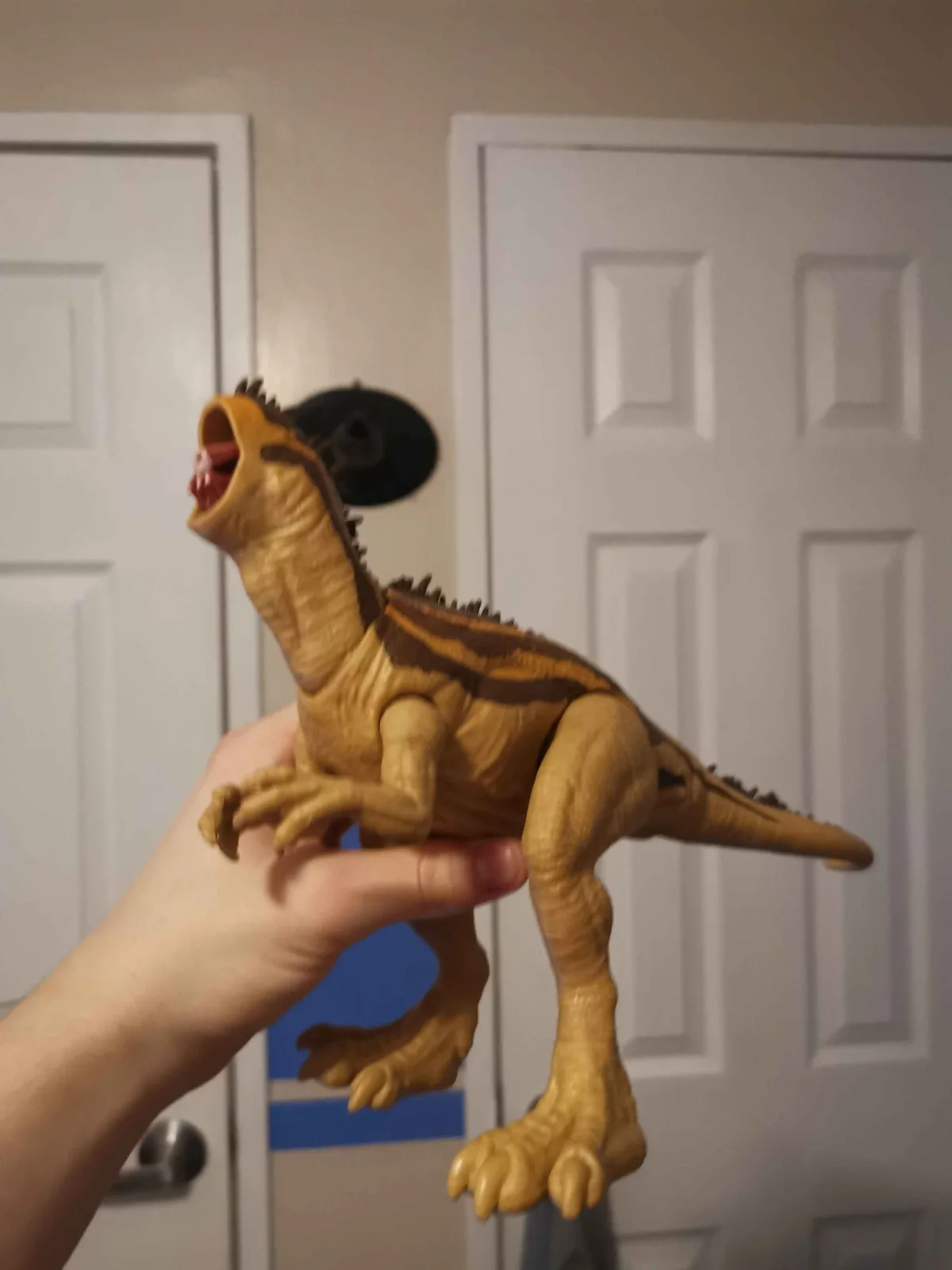 A person holds a tan and brown toy dinosaur with its mouth open, positioned in front of two white doors. The dinosaur has a…