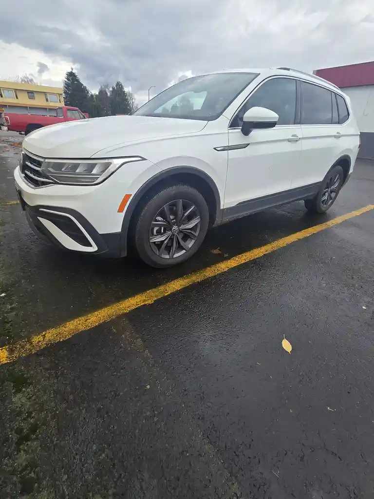 A white Volkswagen Tiguan SUV is parked at an angle in a wet parking lot, partially over a yellow line, with a red truck and…