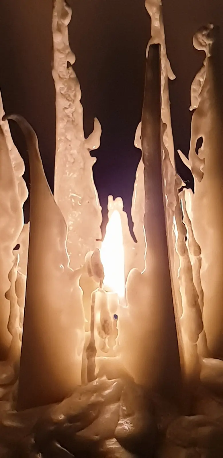 A close-up photo shows several tall, thick candles covered in thick, dripping wax that has solidified into icicle-like…