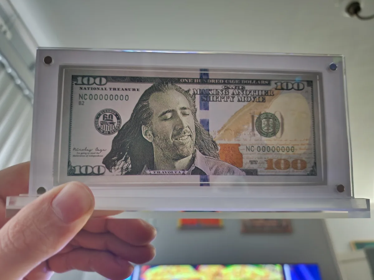 A hand holds a clear acrylic display case containing a fake $100 bill. The bill features Nicolas Cage's face, with…