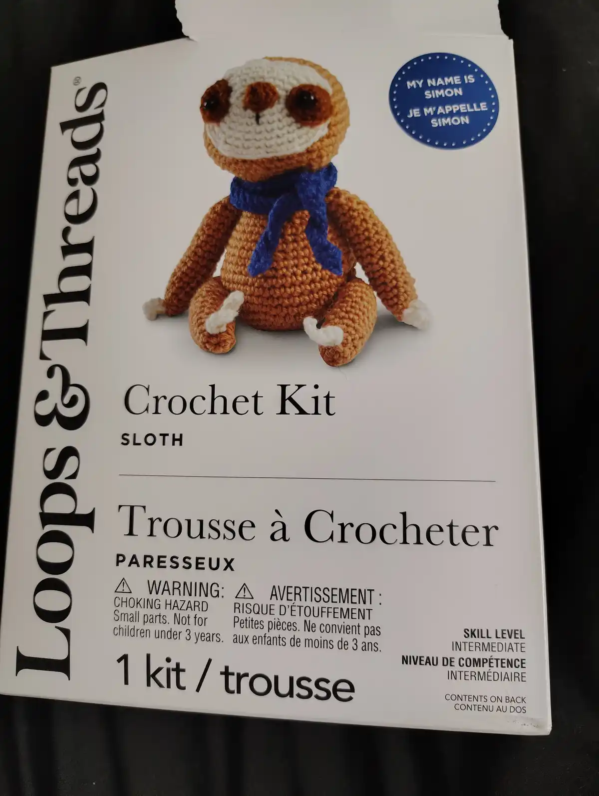 A white box for a 'Loops & Threads' crochet kit featuring a brown and white crocheted sloth wearing a blue scarf. The sloth…