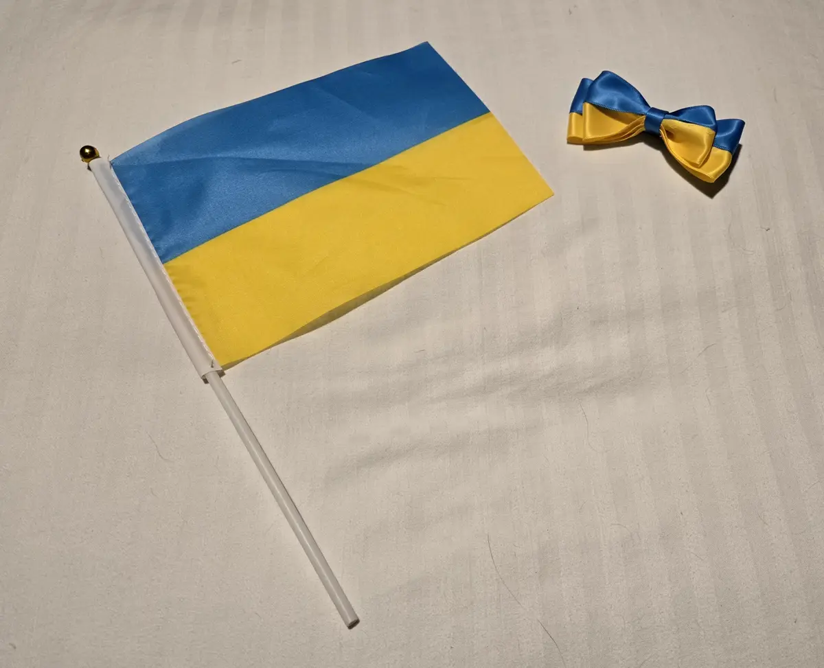 A small Ukrainian flag on a white stick with a gold tip lies next to a matching blue and yellow bow tie on a light gray…