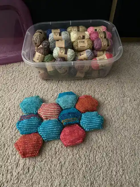 A clear plastic bin filled with colorful yarn skeins sits on a carpet, with a small pile of knitted hexagonal pieces…
