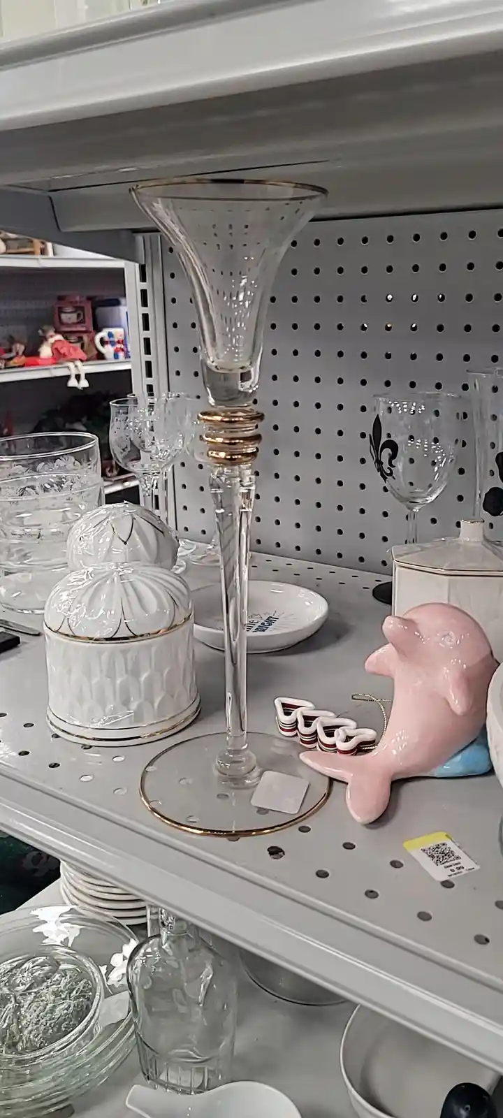A tall, ornate glass candlestick with gold trim stands on a retail shelf surrounded by various glassware and ceramic items…