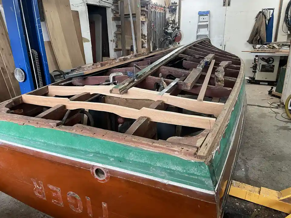 A wooden boat named 'TIGER' is shown in a workshop, partially disassembled with its interior ribs exposed and held together…