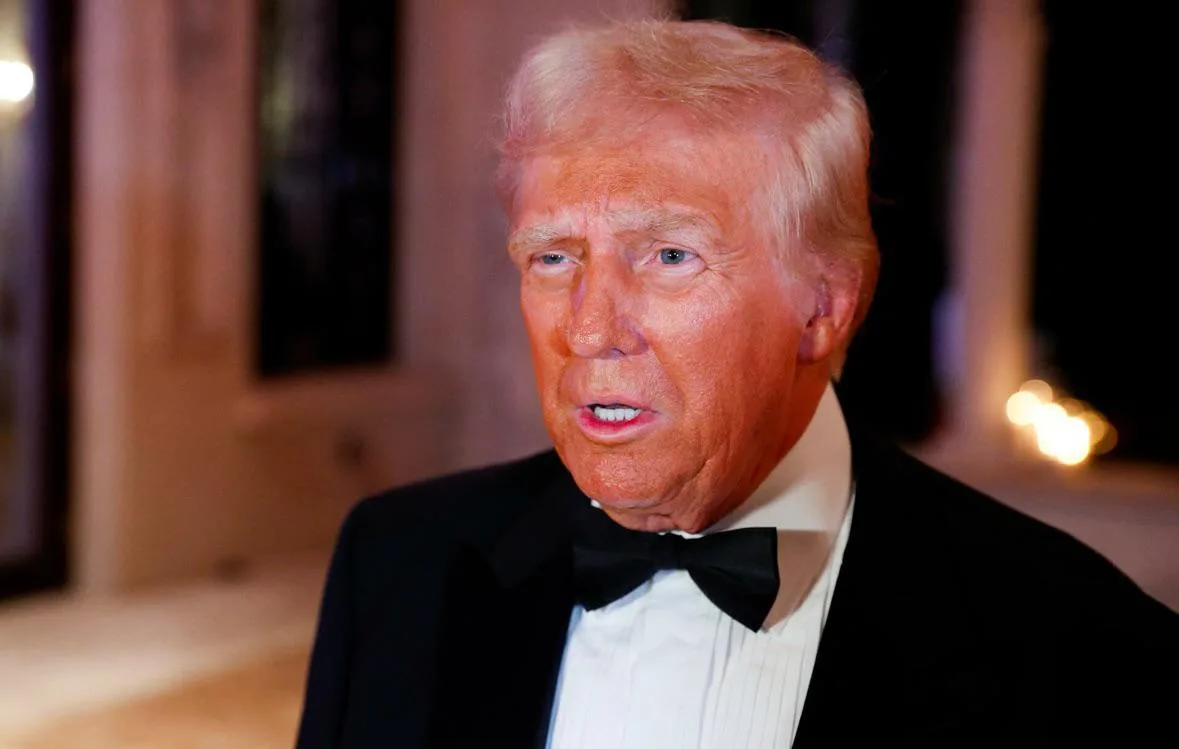 a close-up photo of Donald Trump wearing a black tuxedo and bow tie, looking slightly to his right with a serious…