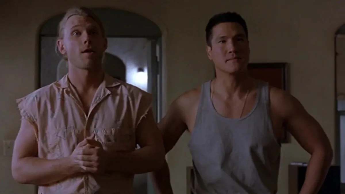two men stand in a dimly lit room; the man on the left, wearing a pink sleeveless shirt, looks upward with a shocked…