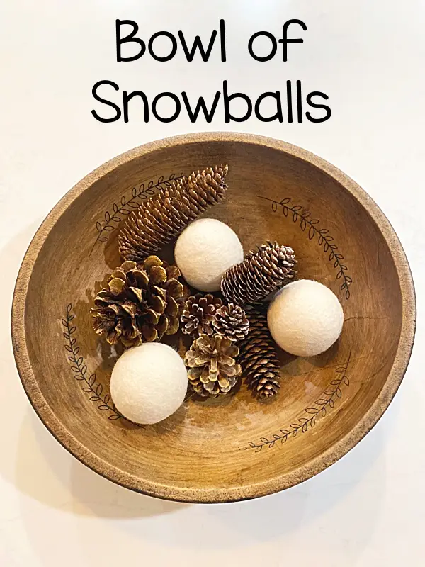 A wooden bowl with decorative leaf patterns contains several pinecones and three white, round felt balls that resemble…