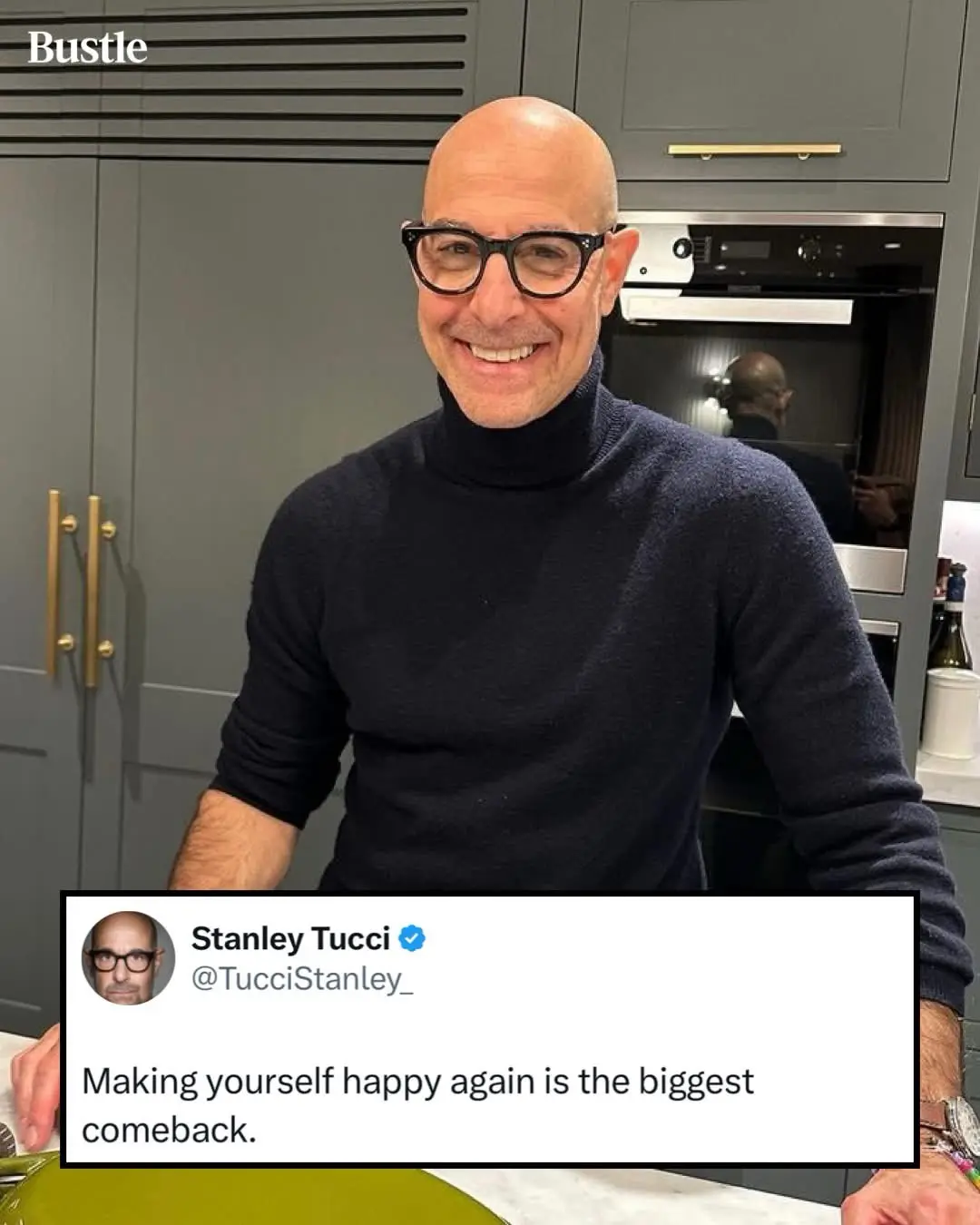 A bald man with black-rimmed glasses and a dark turtleneck sweater smiles broadly in a modern kitchen with grey cabinets and…