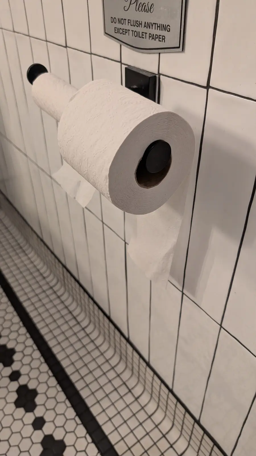 A close-up photo shows a toilet paper roll on a black holder mounted on a white tiled wall. Above the roll, a sign reads…