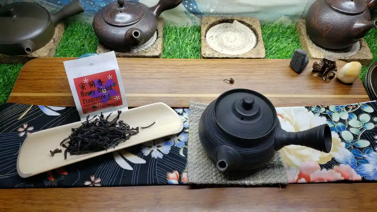 A still life of a traditional tea setup featuring a black ceramic teapot on a woven mat, next to a bamboo tray holding dark…