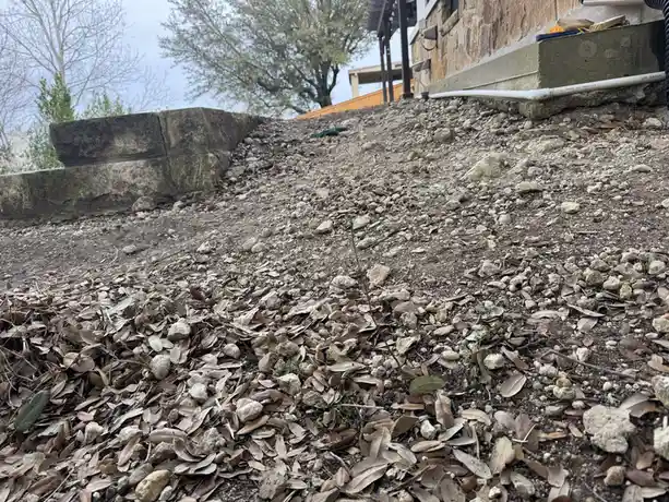 A low-angle photo of a steep, rocky, leaf-covered hillside next to a stone retaining wall and house foundation. The overlaid…