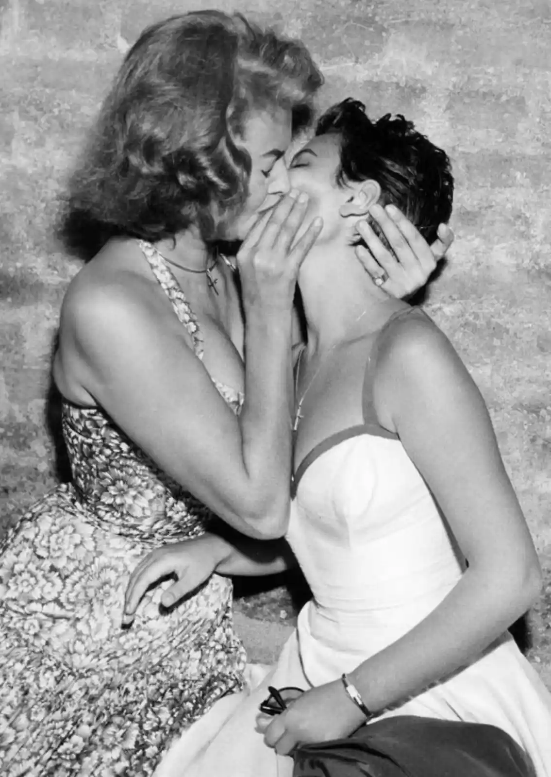 A vintage black-and-white photo shows two women in dresses kissing passionately; one woman kisses the other on the cheek…