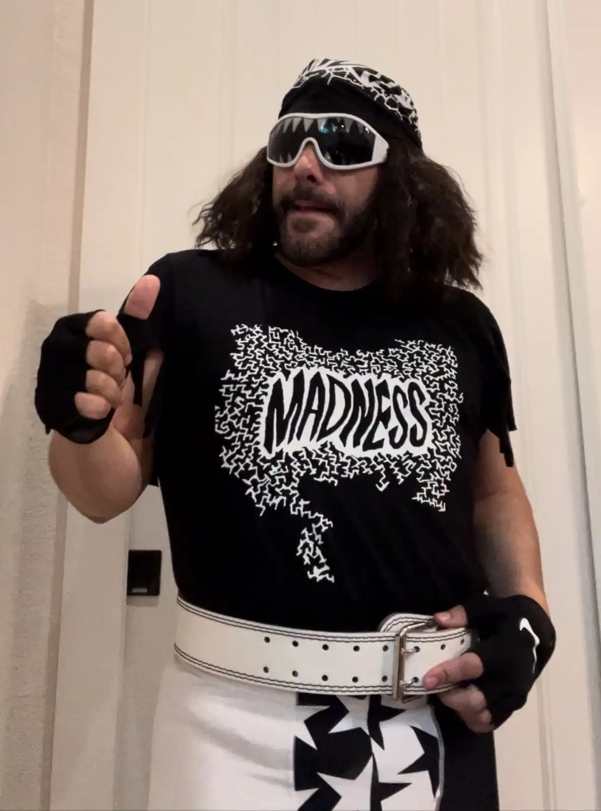 A man with long dark hair, a beard, and wearing a black t-shirt with the word 'MADNESS' in a jagged white font, white…