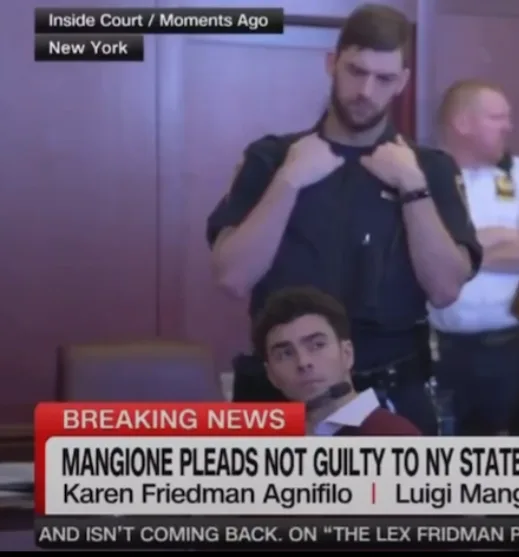 A man in a courtroom sits with a shocked expression as a police officer behind him adjusts his shirt. A news banner reads…