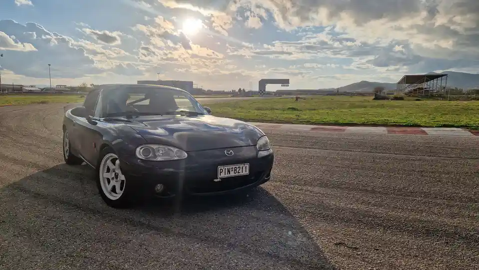 A dark blue Mazda Miata convertible is parked on an asphalt racetrack under a partly cloudy sky with the sun shining. The…