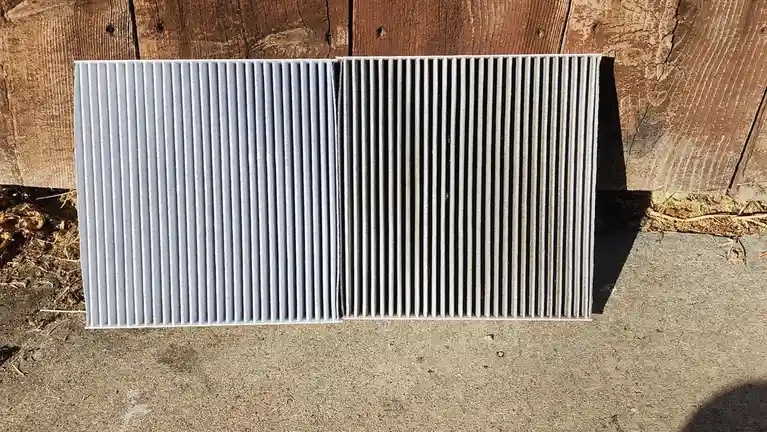 Two side-by-side pleated air filters lie on a concrete surface against a wooden fence; the left filter is visibly clean and…