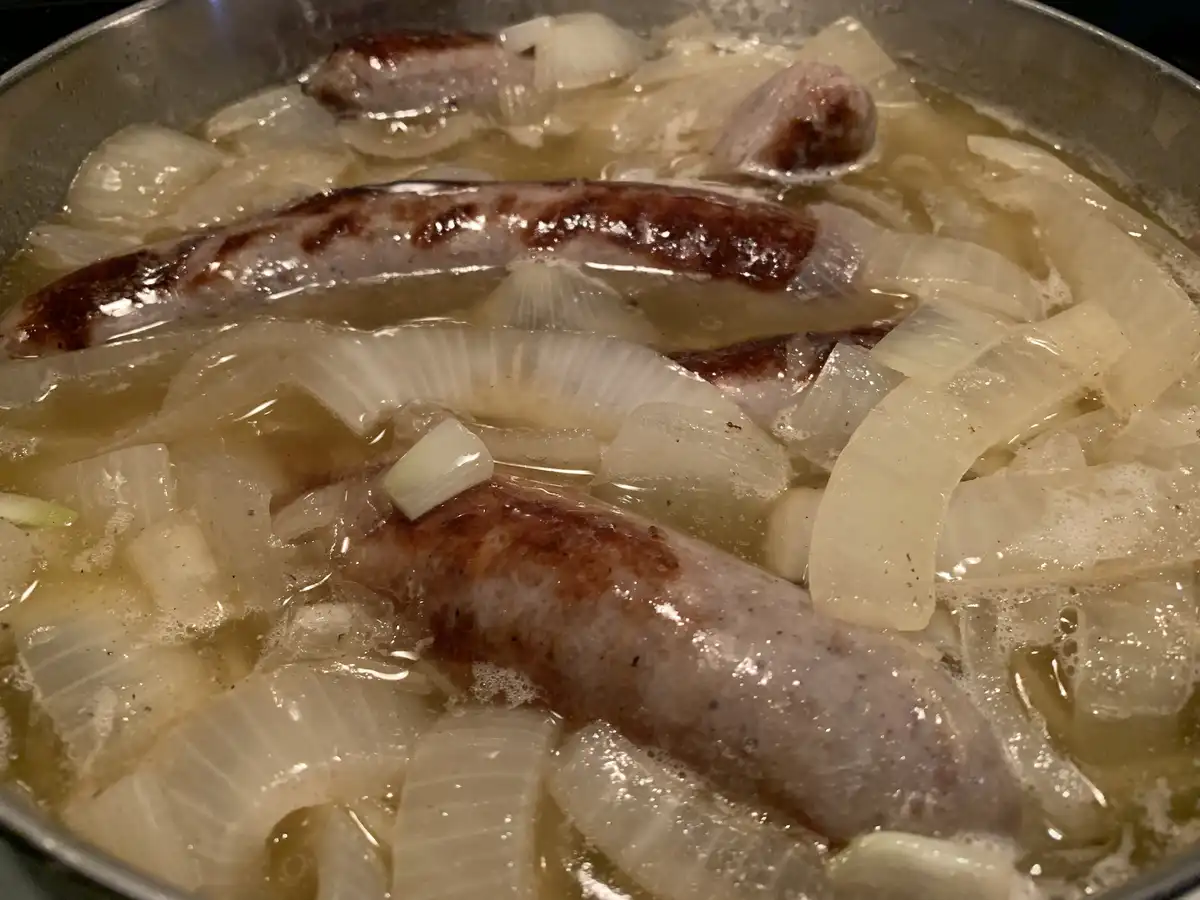 A close-up photo shows several sausages and sliced onions simmering in a pan with bubbling liquid. The sausages are…