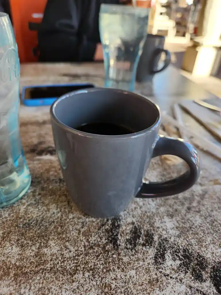 A close-up photo shows a dark grey coffee mug filled with black coffee sitting on a speckled countertop. Beside it are a…