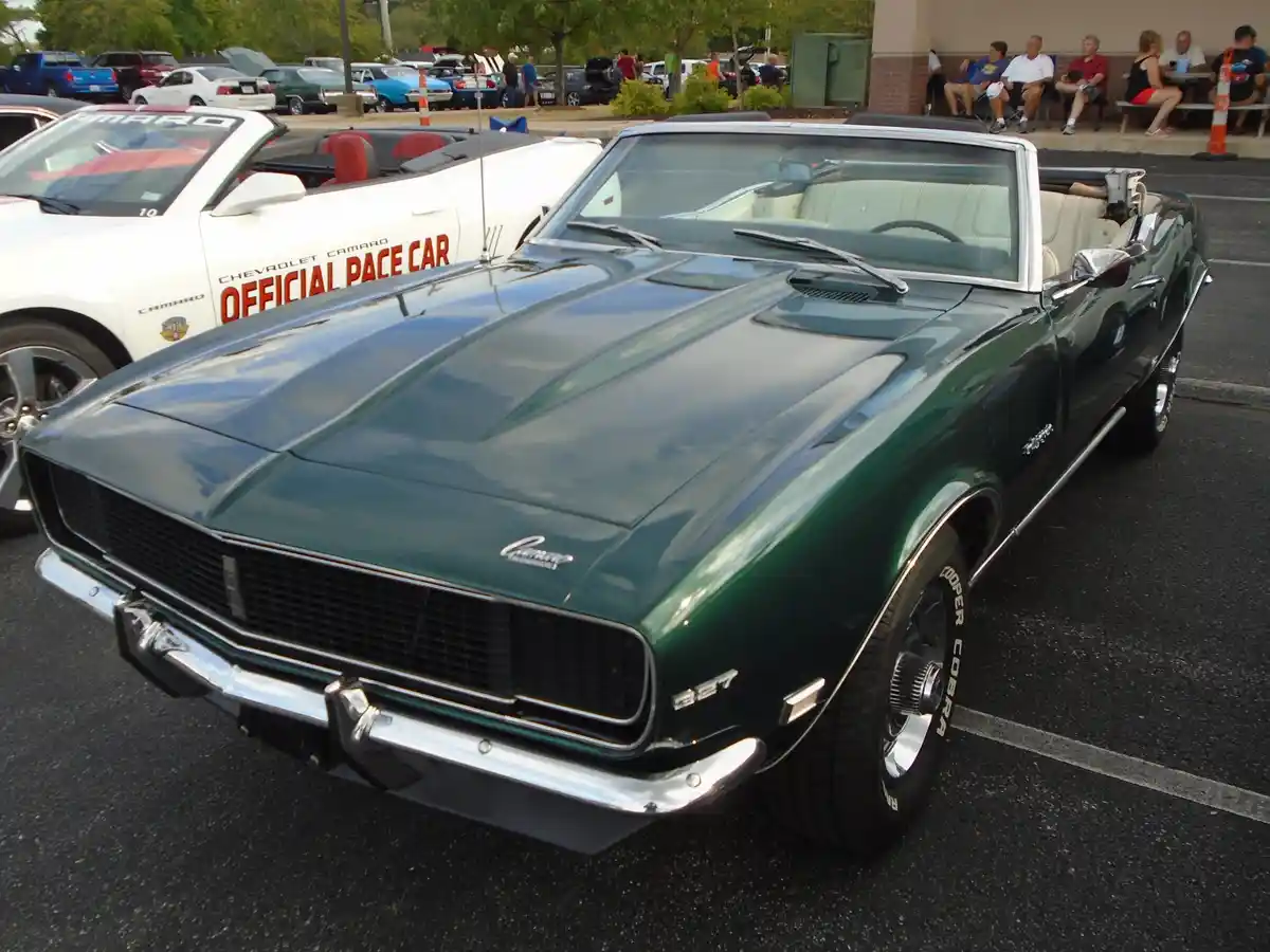 A green 1967 Chevrolet Camaro convertible is parked in a lot, with a white Camaro pace car visible behind it. The green car…