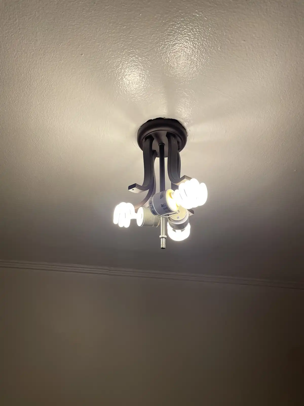 A photo of a ceiling light fixture with three lit compact fluorescent bulbs, one of which is visibly broken and hanging by a…