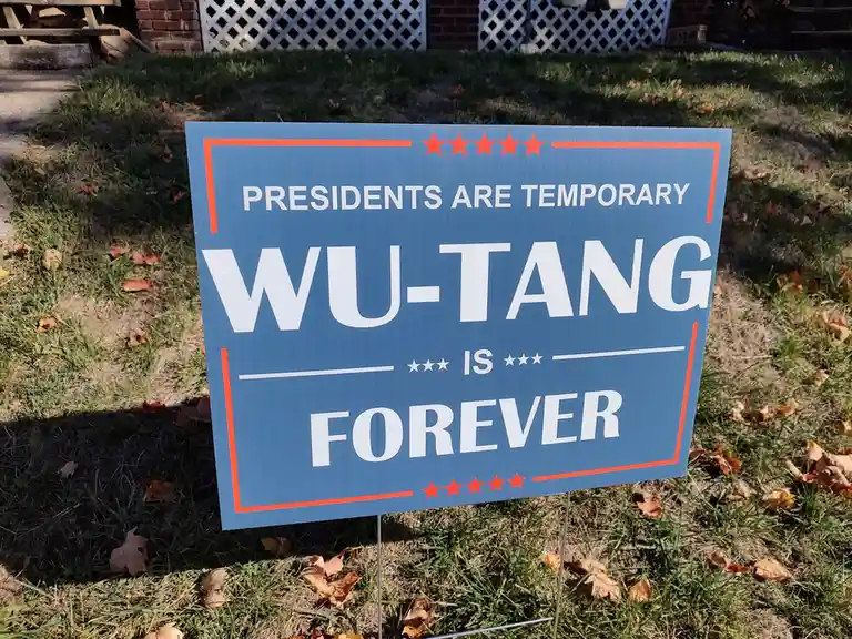 A blue political-style yard sign with orange borders and red stars, planted in grass with fallen leaves, reads 'PRESIDENTS…