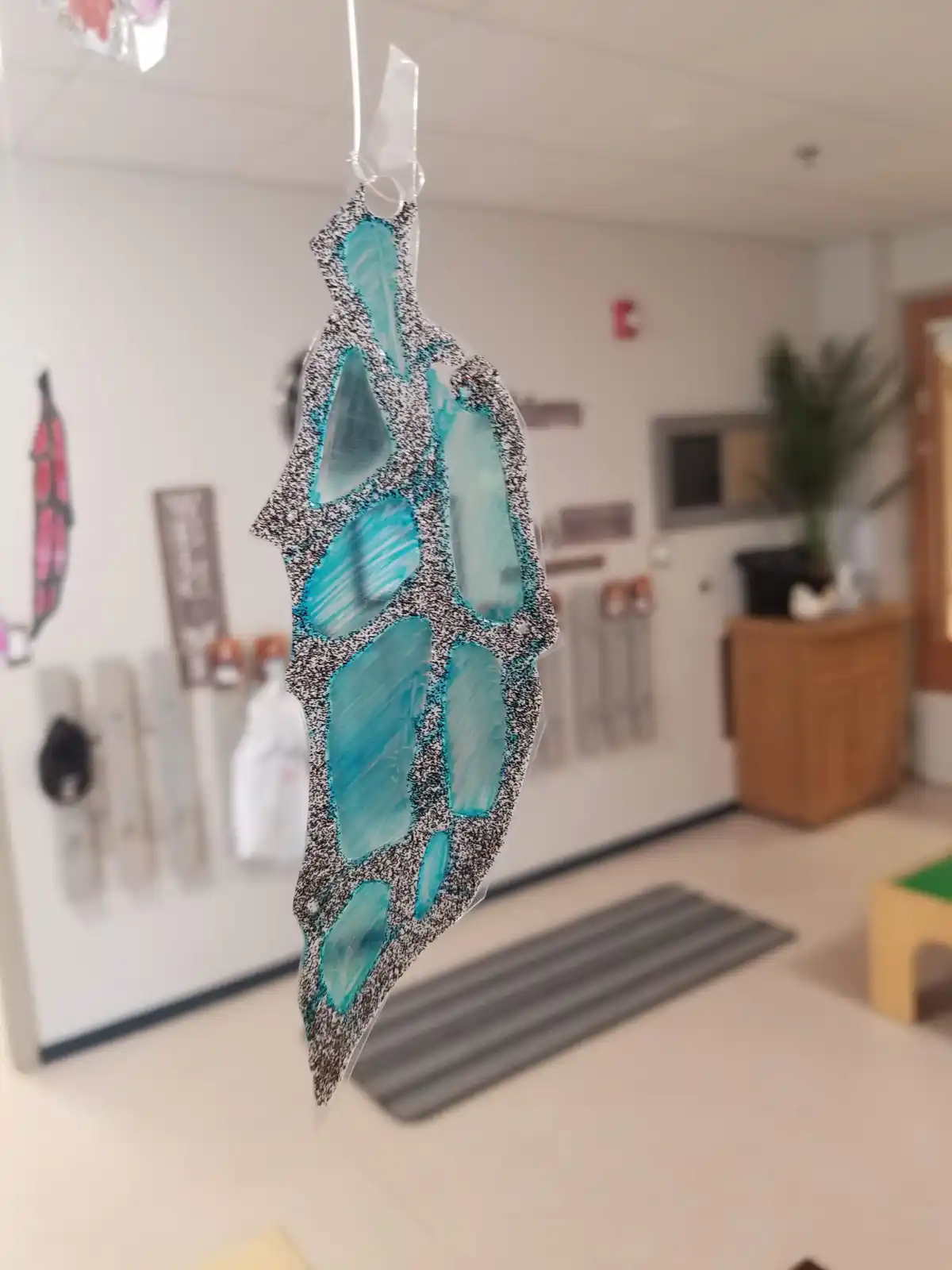 A handmade feather-shaped craft, likely made from a decorated notebook page with blue paint sections, hangs from a clear…