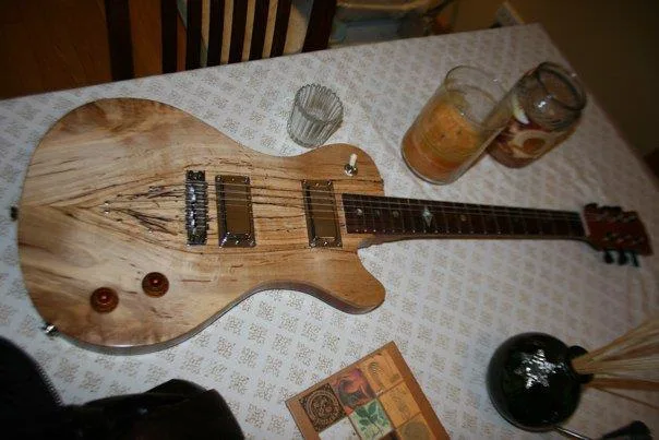 A light-colored, wood-grain electric guitar with a natural finish lies on a patterned tablecloth, surrounded by glasses, a…