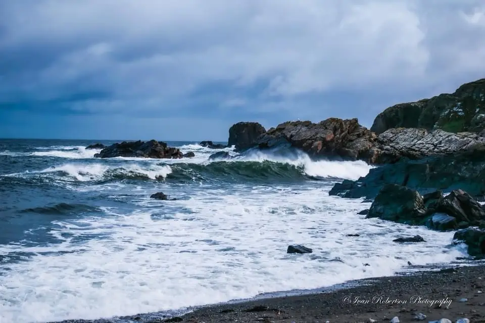 A dramatic photograph of turbulent ocean waves crashing against dark, jagged rocks under a stormy, cloudy sky. The…