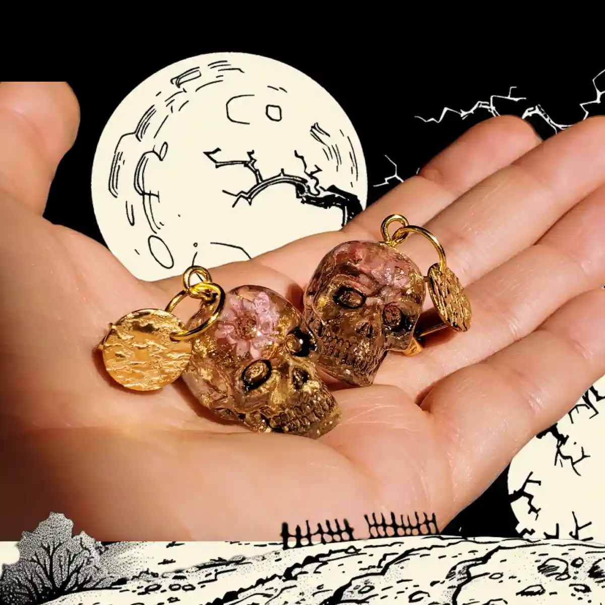 A hand holds two small, gold-colored skull charms embedded with pink resin, each attached to a gold chain and a small gold…