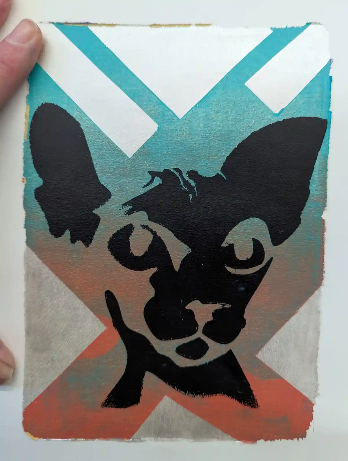 A hand holds a painted artwork featuring a black silhouette of a dog's face with large ears and a pointed snout, set against…