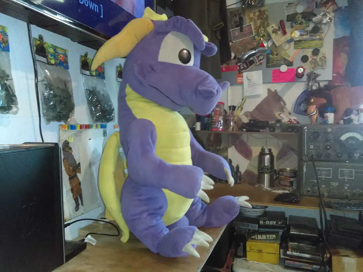 A large purple and yellow plush dragon toy, resembling Spyro the Dragon, is sitting upright on a cluttered desk in a messy…