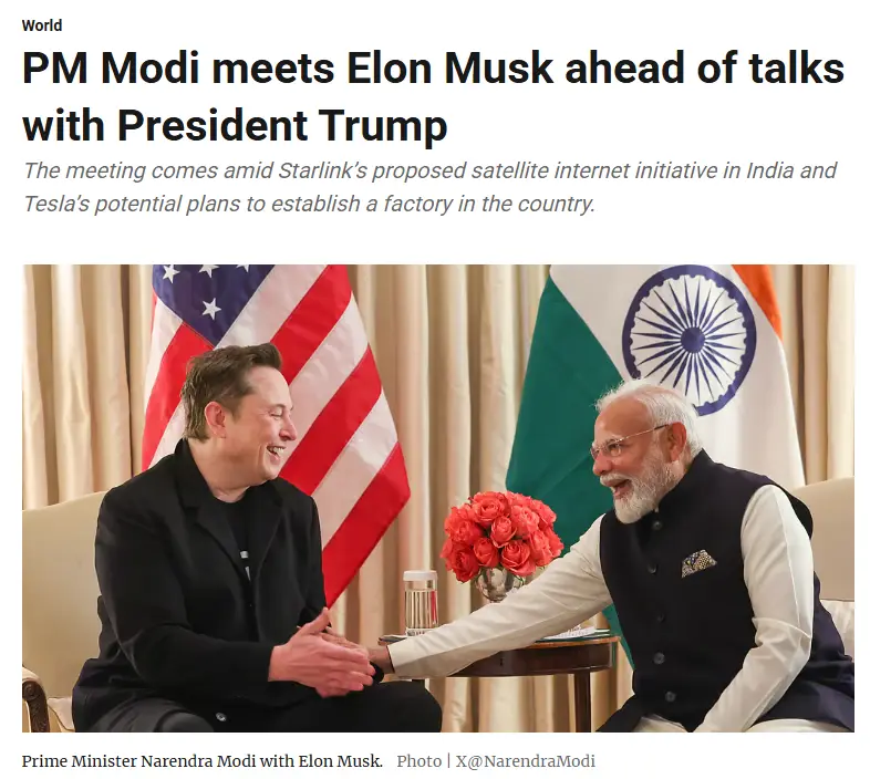 Two men are seated and shaking hands in front of American and Indian flags. The man on the left, wearing a black jacket, is…