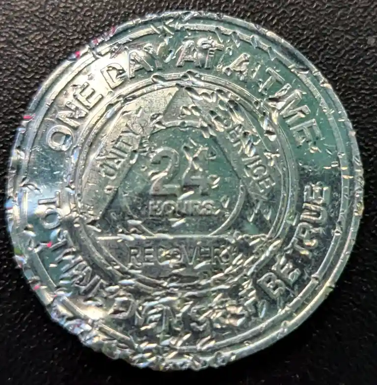 A close-up of a worn, circular metal token or medallion with embossed text reading 'ONE DAY AT A TIME' around the outer…