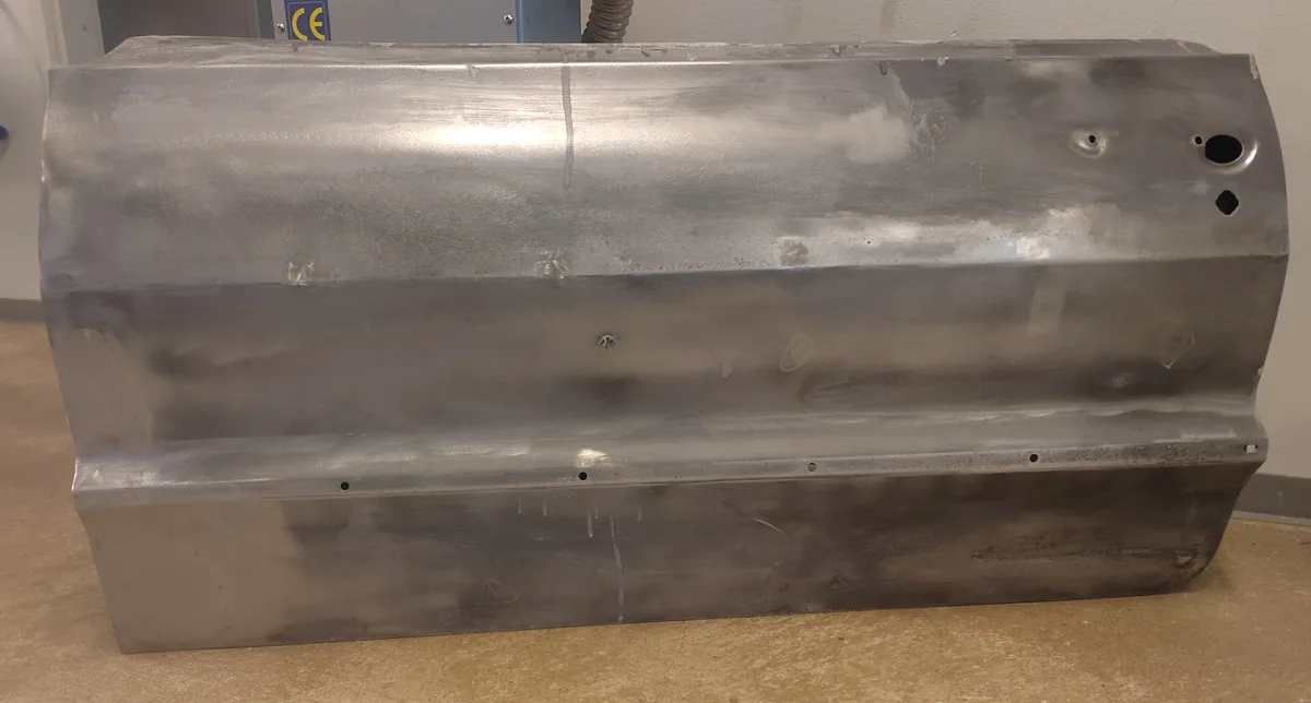 A large, shiny, silver metal automotive part, possibly a fender or bumper, is photographed upright on a tan floor. It has a…