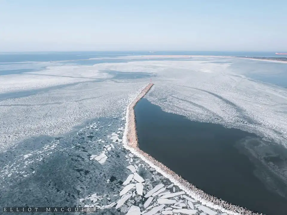 An aerial photo shows a long, narrow stone breakwater extending into icy waters, with a wide expanse of frozen sea…