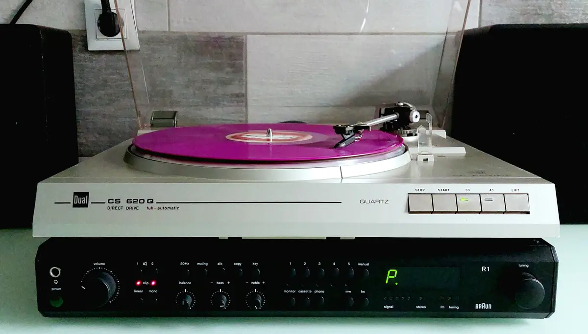A vintage Dual CS 620 Q turntable with a bright pink vinyl record spinning, placed atop a Braun R1 stereo receiver. The…