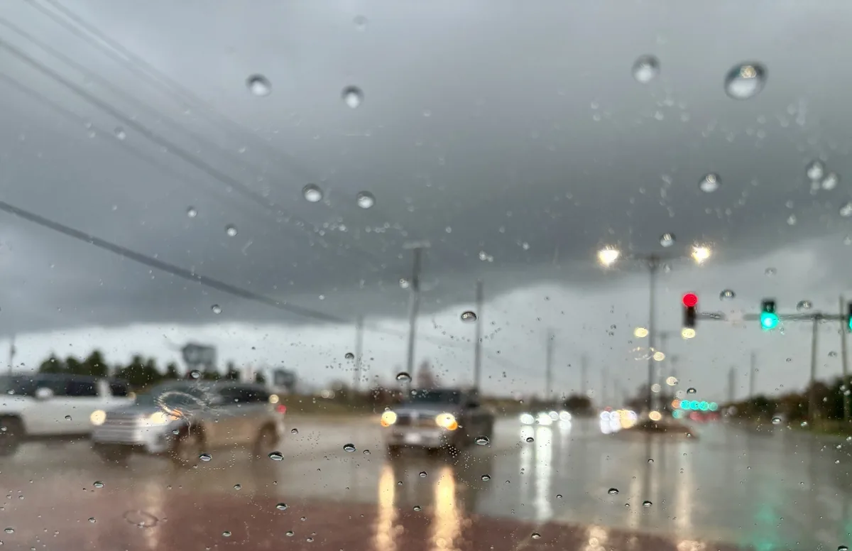 A blurry, rain-streaked view from inside a car shows wet pavement, headlights of other vehicles, and traffic lights, with…