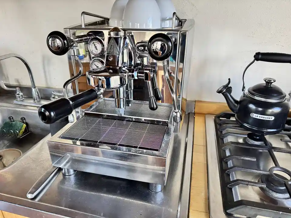 A shiny, professional-grade espresso machine sits on a stainless steel countertop next to a gas stove with a black Le…