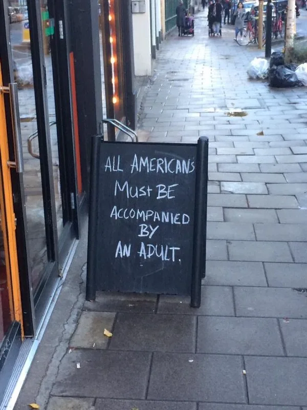 A black chalkboard sign on a sidewalk outside a storefront reads 'ALL AMERICANS Must BE ACCOMPANIED BY AN ADULT.' The sign…