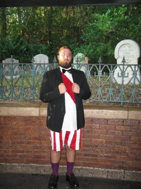 A man with a beard and glasses stands in a cemetery wearing a black suit jacket, white shirt, black bowtie, red sash…