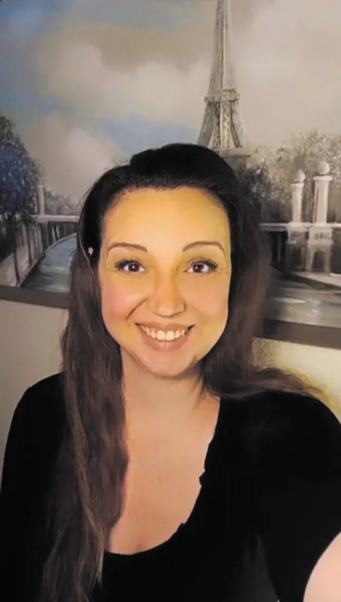 A woman with long dark hair and a black top smiles at the camera while standing in front of a painted backdrop featuring the…