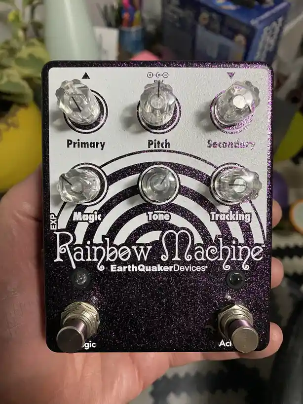 A hand holds a purple glitter-finished guitar effects pedal labeled 'Rainbow Machine' by EarthQuakerDevices, featuring knobs…