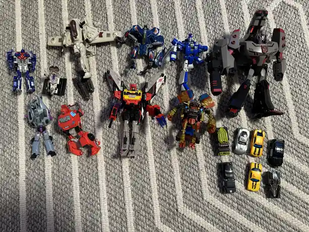 An overhead shot displays a collection of various Transformers action figures and their corresponding vehicle modes arranged…